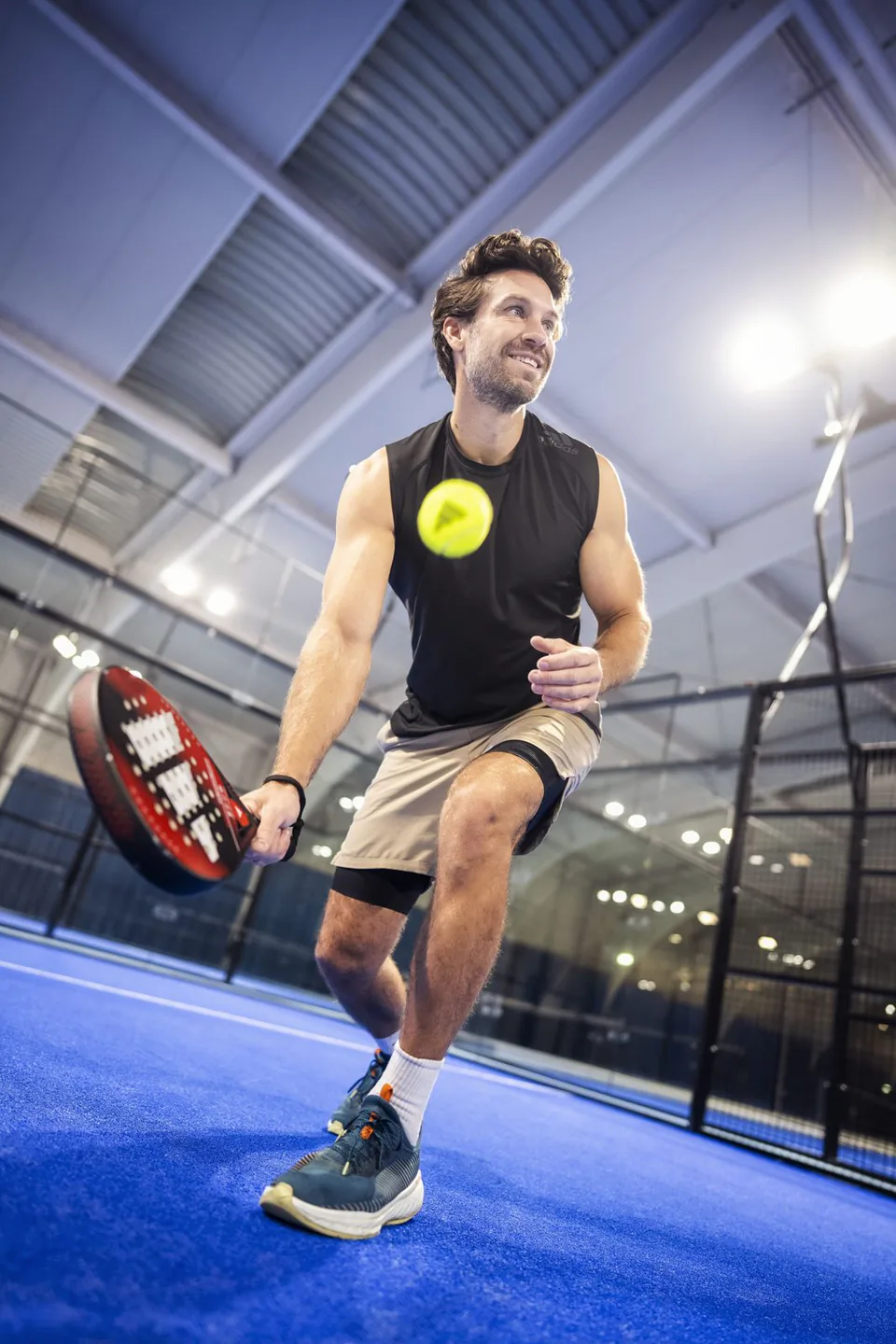 Mann mit Padelschläger spielt einen gelben Ball auf blauem Hallenplatz unter hellen Deckenleuchten.